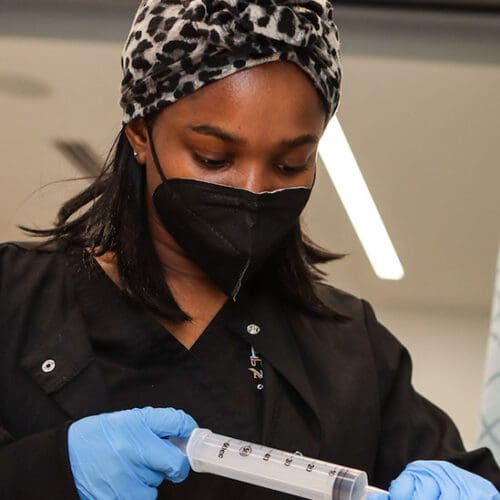 A nursing student uses a syringe during a lab. They are wearing black scrubs, blue gloves, a black face mask and a cheetah print hair wrap.