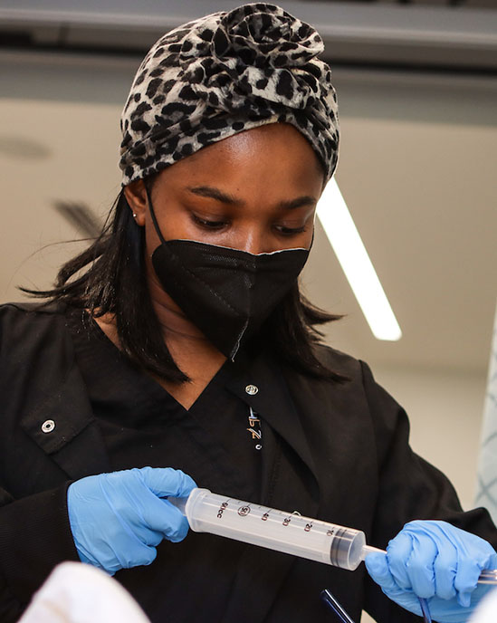 A nursing student uses a syringe during a lab. They are wearing black scrubs, blue gloves, a black face mask and a cheetah print hair wrap.
