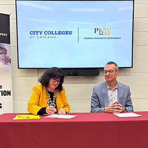 A woman and man in professional clothing sign an agreement on a table in front of a screen with logos for the City Colleges of Chicago and PNW