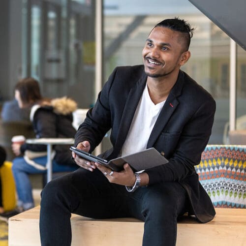 A student sits on a couch. He is looking off to the left of the camera. There is a folder in his hands.