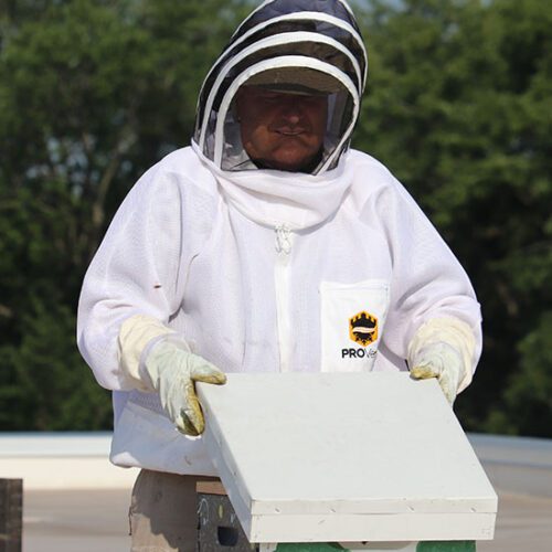John Bachmann stands in a beekeeper suit. He is lifting the lid off of a beehive.