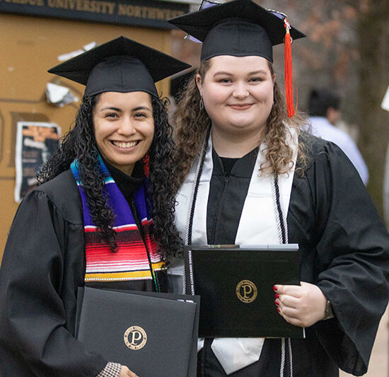 Two students in their cap and gown pose together outside and hold up their degree holders