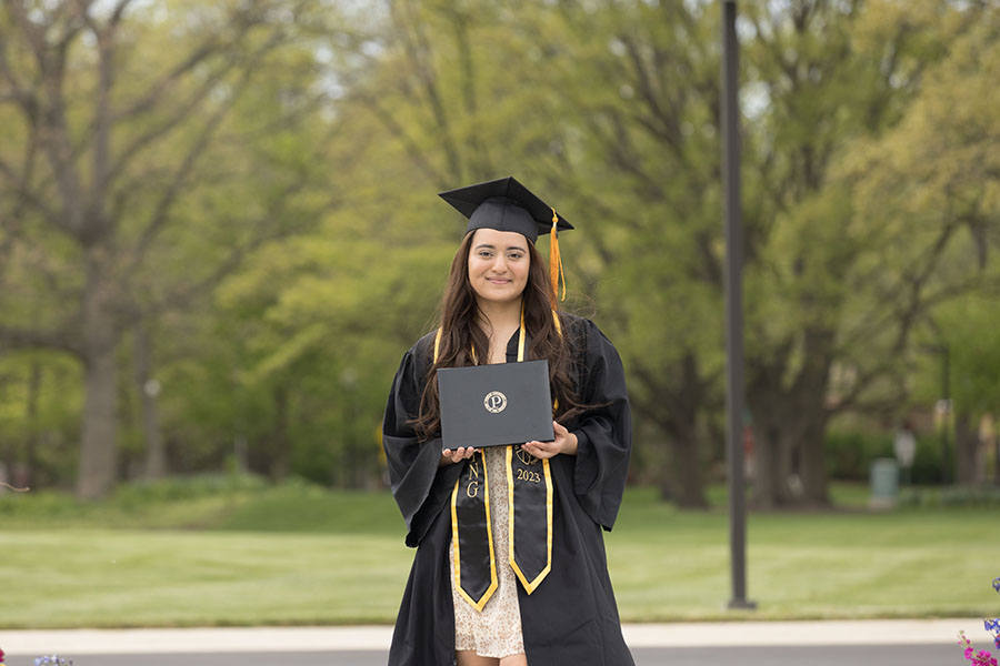 A student stands in black graduation regalia. They are holding up their degree frame