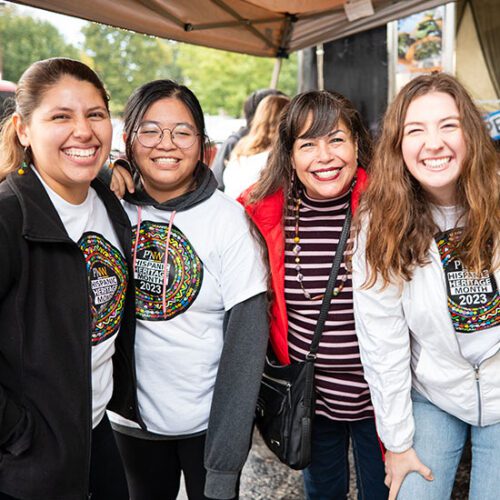 Four people lean into each other and pose. They are all smiling. Three of the people are wearing PNW's hispanic heritage month shirts while the fourth person is wearing a striped shirt.