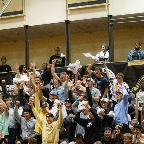 Crowd of students sit on indoor gymnasium bleachers as they cheer on athletics event.