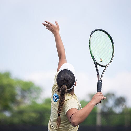 A PNW womens tennis player throws a ball into the air to serve. She is holding a tennis racket.