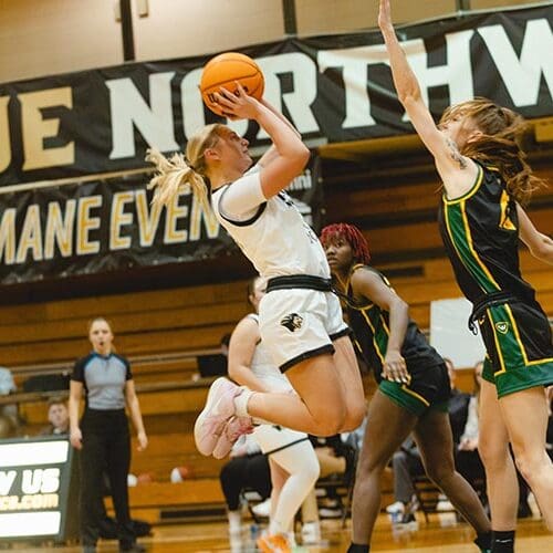 Women's Basketball photo. Player shooting the basketball while opposing team tries blocking it.