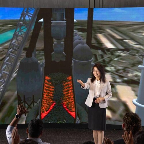 A woman stands in front of a visualization of a steelmaking blast furnace at Purdue University Northwest.