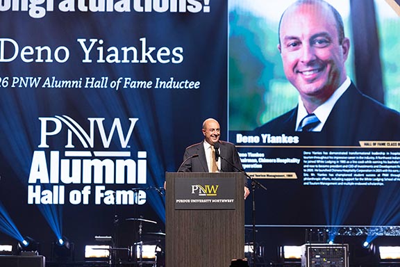 Deno Yiankes stands at a podium, information about his Hall of Fame award is on the screen behind him.