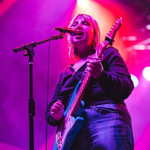 Lili Trifilio, the lead singer of Beach Bunny, stands in front of a microphone and sings while playing the guitar