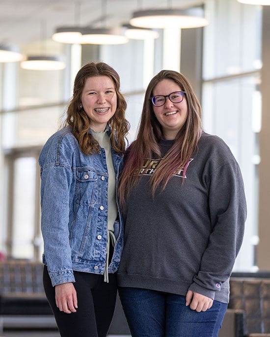 Two students stand together in a hallway. They are smiling and each have one arm wrapped around the other