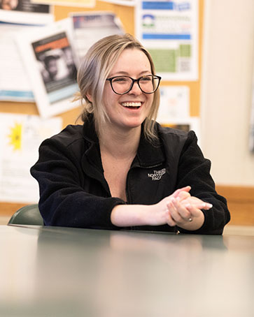 A student sits at a table, smiling. Their hands are folded and sitting on the table.