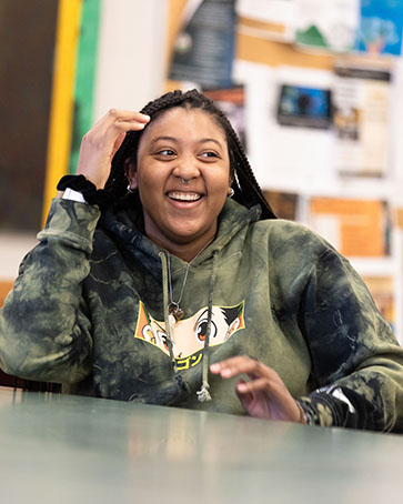 A student sits at a table and smiles. They have one hand in their hair and one hand on the table.