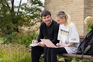 Two PNW students sitting on a bench outside while working on a laptop and talking 363x242