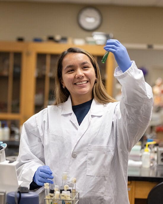 A student in a white lab coat holds a vile containing green liquid in the air