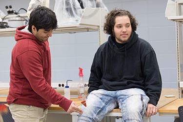 A student in a black hoodie and jeans sits on a table. A student in a red hoodie and khakis stands next to the table and checks the other student's knee reflexes.