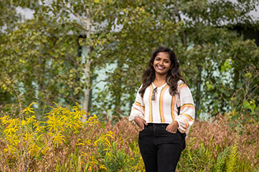 A student stands in front of foliage. They are smiling with their hands in their pocket.
