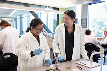 two students in lab coats work together on a burner in a biology lab