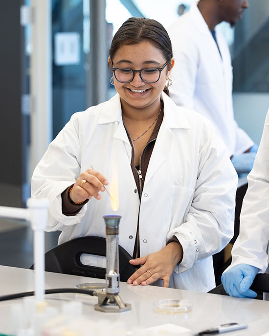 A student in a labcoat holds an item over a burner.