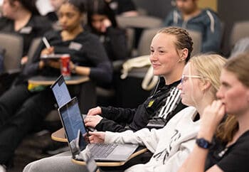 Students sit in a lecture room with laptops open on the desks in front of them.