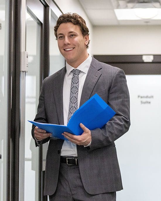 A student stands in a hallway. He is wearing a suit and holding a blue folder.