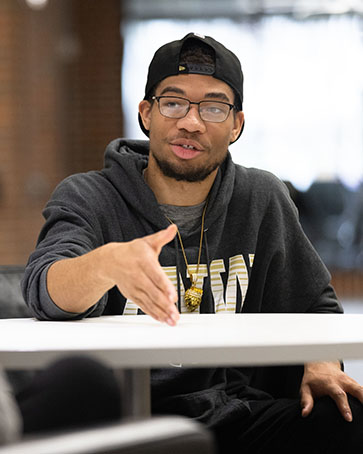 A student sits at a table. He is wearing a PNW hoodie and backwards baseball cap