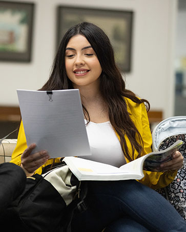 A student in a yellow blazer sits on a couch. There is a notebook on her lap and she is holding a stack of papers that are held together by a binder clip