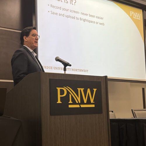David Pratt stands behind a wooden podium with the "PNW" logo on it. He is speaking into a microphone. A slideshow for his presentation is being projected onto the screen behind him