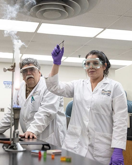 Student creates a chemical reaction. There is some smoke coming out of a test tube on the table in front of them. There is also a professor in the photo
