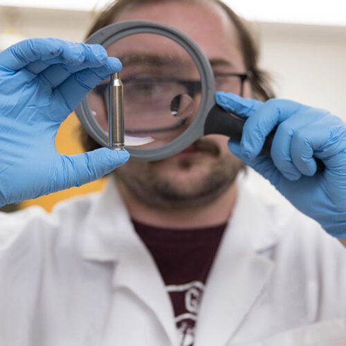 A PNW student in a lab coat looks at a bullet through a magnifying glass.