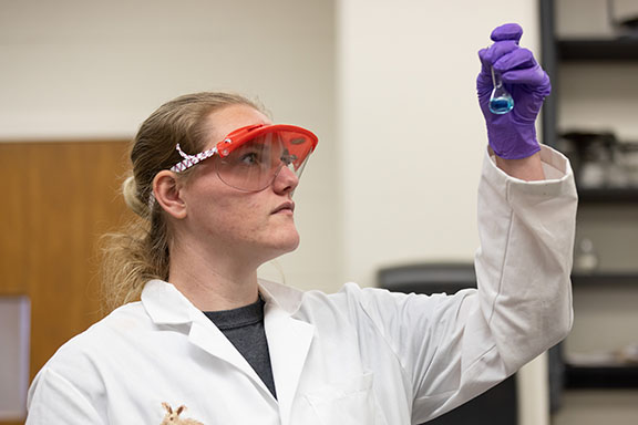 A student in protective goggles, white lab coat, and purple holds a test tube in the air