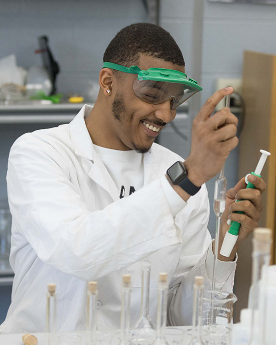 A student pipes liquid into a test tube. They are wearing a white lab coat and green safety goggles.