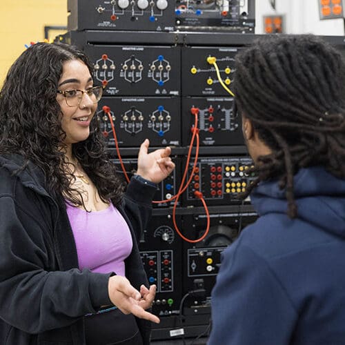 Two students work in an engineering lab. One student is facing the camera and gesturing toward the equipment. The other student is sitting, facing away from the camera.
