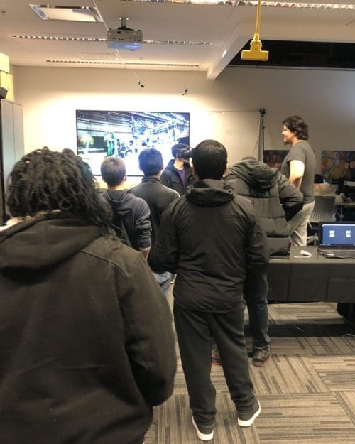 Visitor participates in hands-on experiences of AR & VR projects in the visualization lab. Participant is standing while wearing a tethered VR headset and manipulating hand held controllers surrounded by several people observing