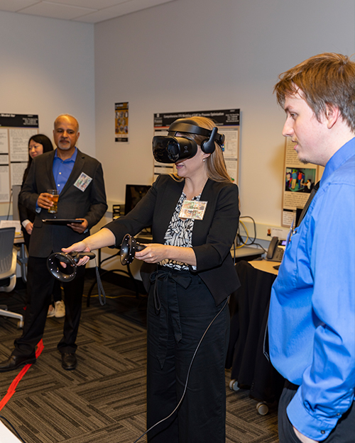 Visitor participates in hands-on experiences of AR & VR projects in the visualization lab. Participant is standing while wearing a tethered VR headset and manipulating hand held controllers surrounded by several people observing