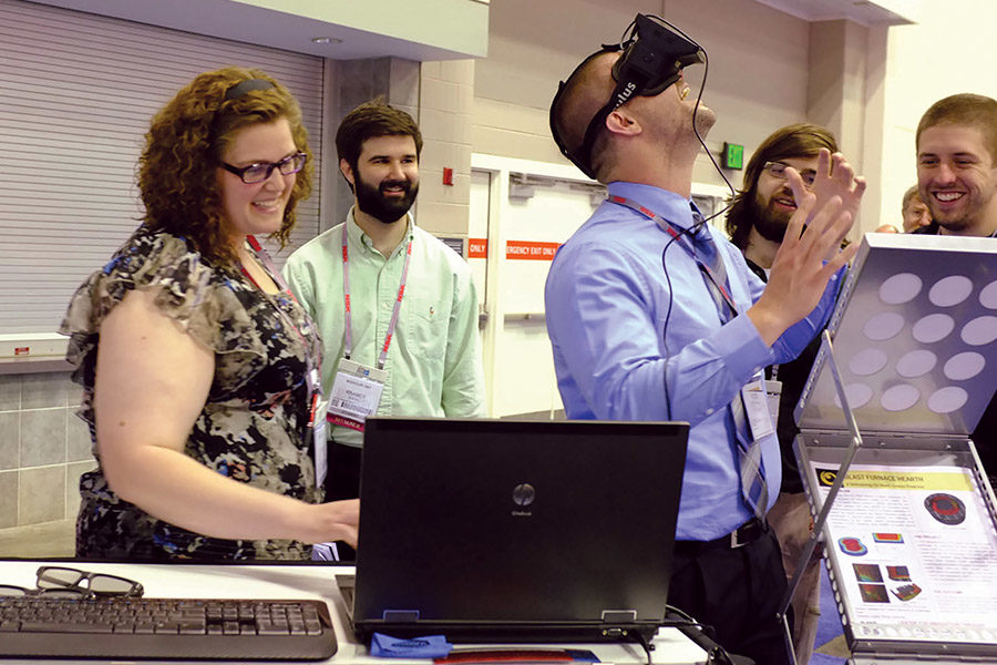 Visitor participates in hands-on experiences of AR & VR projects. Participant is standing while wearing a tethered VR headset surrounded by several people observing