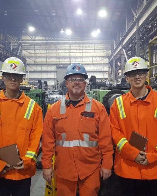 Individuals standing in a row in an industrial setting wearing bright orange uniform jumpsuits and white hard hats and safety goggles