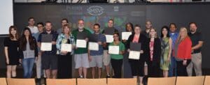 Several people, some holding certificates, standing in a group in front of large screen featuring several logos at the front of an auditorium