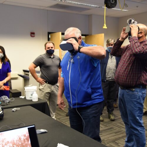 Visitors participate in hands-on experiences of AR & VR projects in the visualization lab. Participants are standing while holding VR goggles surrounded by several people observing