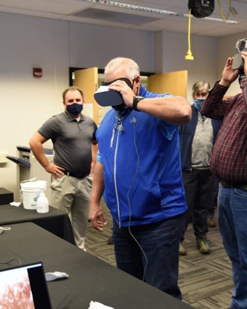 Visitors participate in hands-on experiences of AR & VR projects in the visualization lab. Participants are standing while holding VR goggles surrounded by several people observing