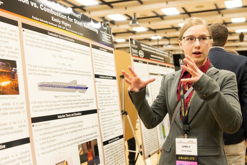 Student standing and talking in front of academic poster in presentation hall with rows of other people and posters in the background