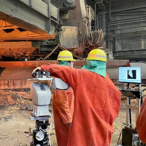 Individuals in an industrial setting wearing bright orange uniform jumpsuits working on standing machinery