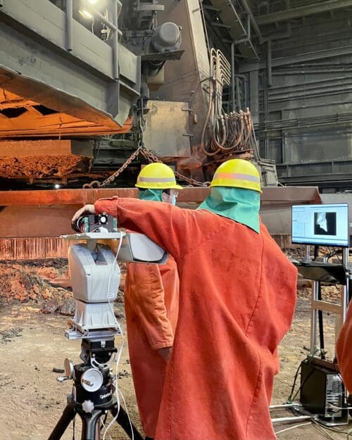 Individuals in an industrial setting wearing bright orange uniform jumpsuits working on standing machinery