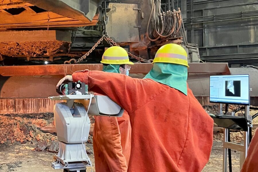 Individuals in an industrial setting wearing bright orange uniform jumpsuits working on standing machinery