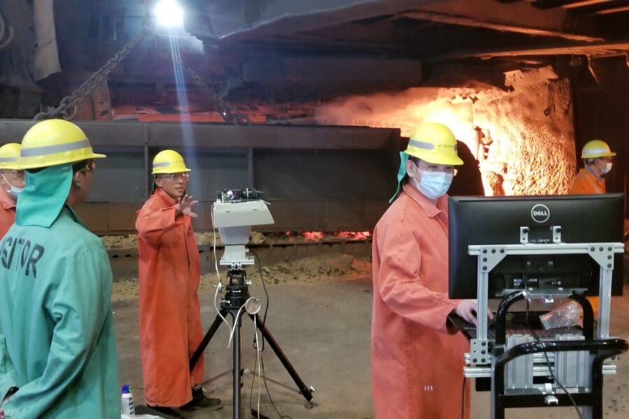 Several individuals in an industrial setting wearing bright orange uniform jumpsuits working on standing machinery