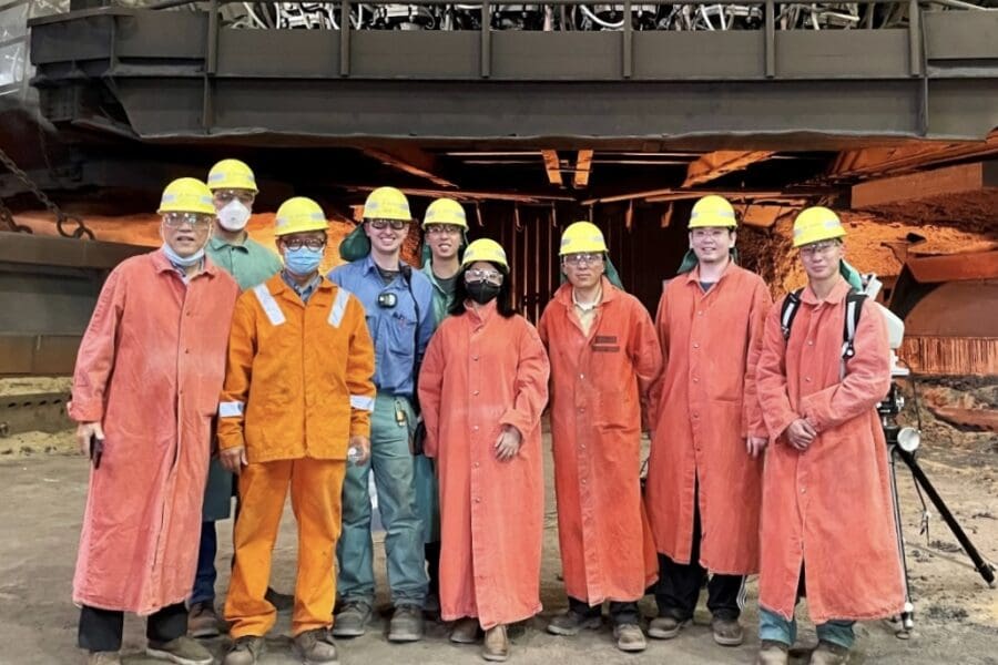 Individuals standing in a row in an industrial setting wearing bright orange uniform jumpsuits and yellow hard hats and safety goggles