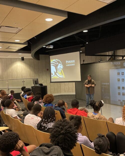 Camp participants sit in chairs watching a presenter standing in front of them.