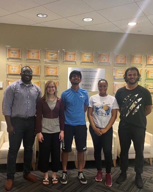 Students in casual attire standing in a row against a patterned wall featuring plaques with several company logos