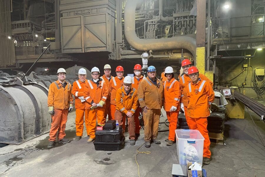 Individuals standing in a row in an industrial setting wearing bright orange uniform jumpsuits and white hard hats and safety goggles