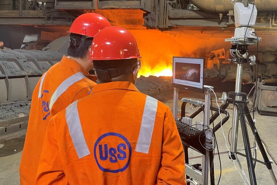 Individuals in an industrial setting wearing bright orange uniform jumpsuits working on standing machinery